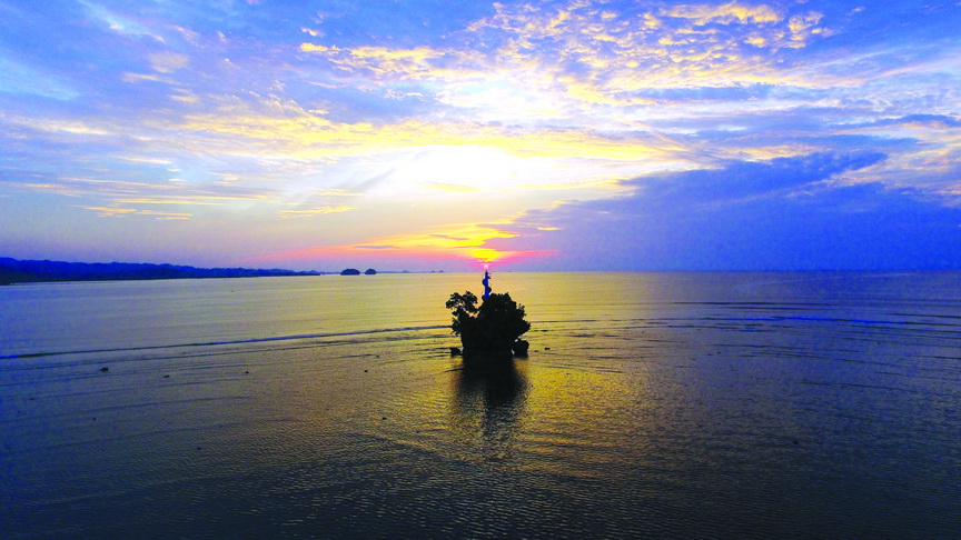 A tree-topped rock islet with a navigational light standing in the waters of Lianga Bay